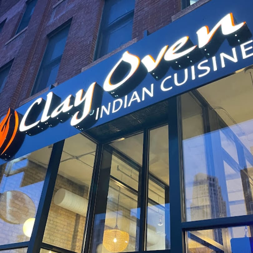 Clay Oven Indian Cuisine restaurant exterior with a blue sign and large windows reflecting a city building.