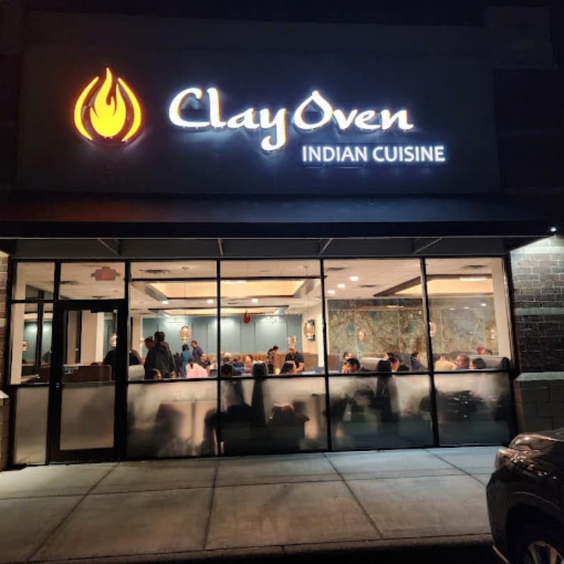 Exterior night view of Clay Oven Indian Cuisine, with a glowing sign and patrons visible inside.