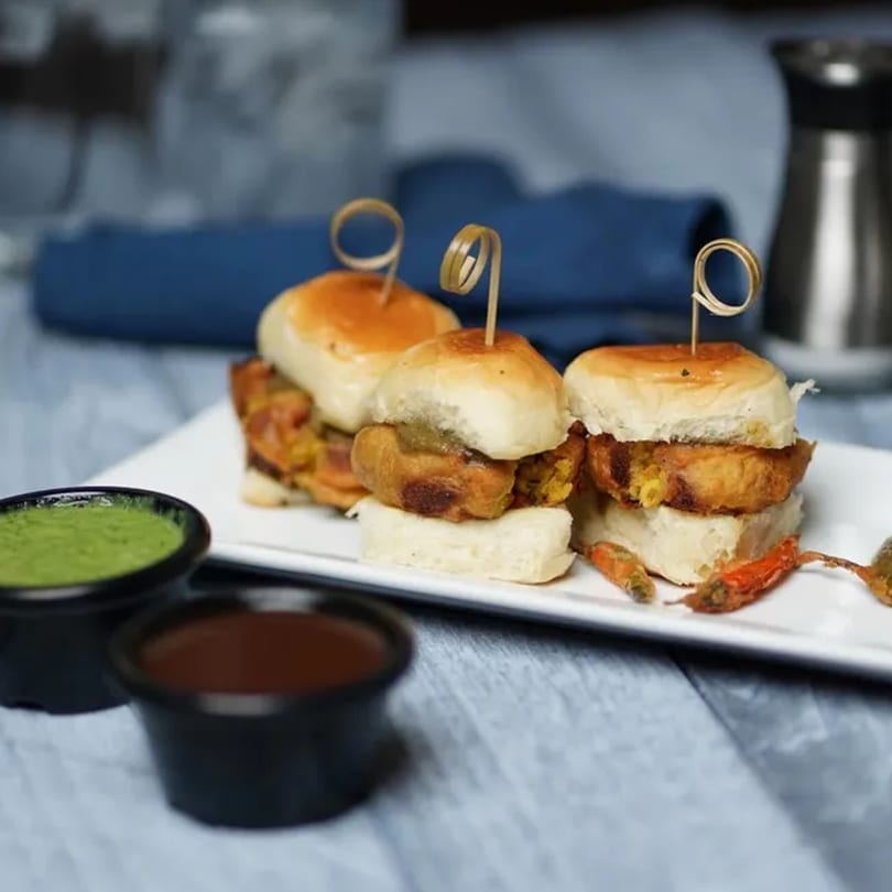 Three Indian vada pav sliders with green and brown dipping sauces on a white plate.