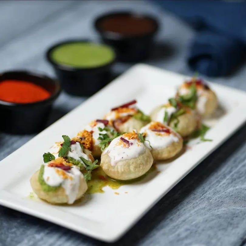 A plate of Indian pani puri, topped with yogurt, spices, and herbs, with dipping sauces in the background.